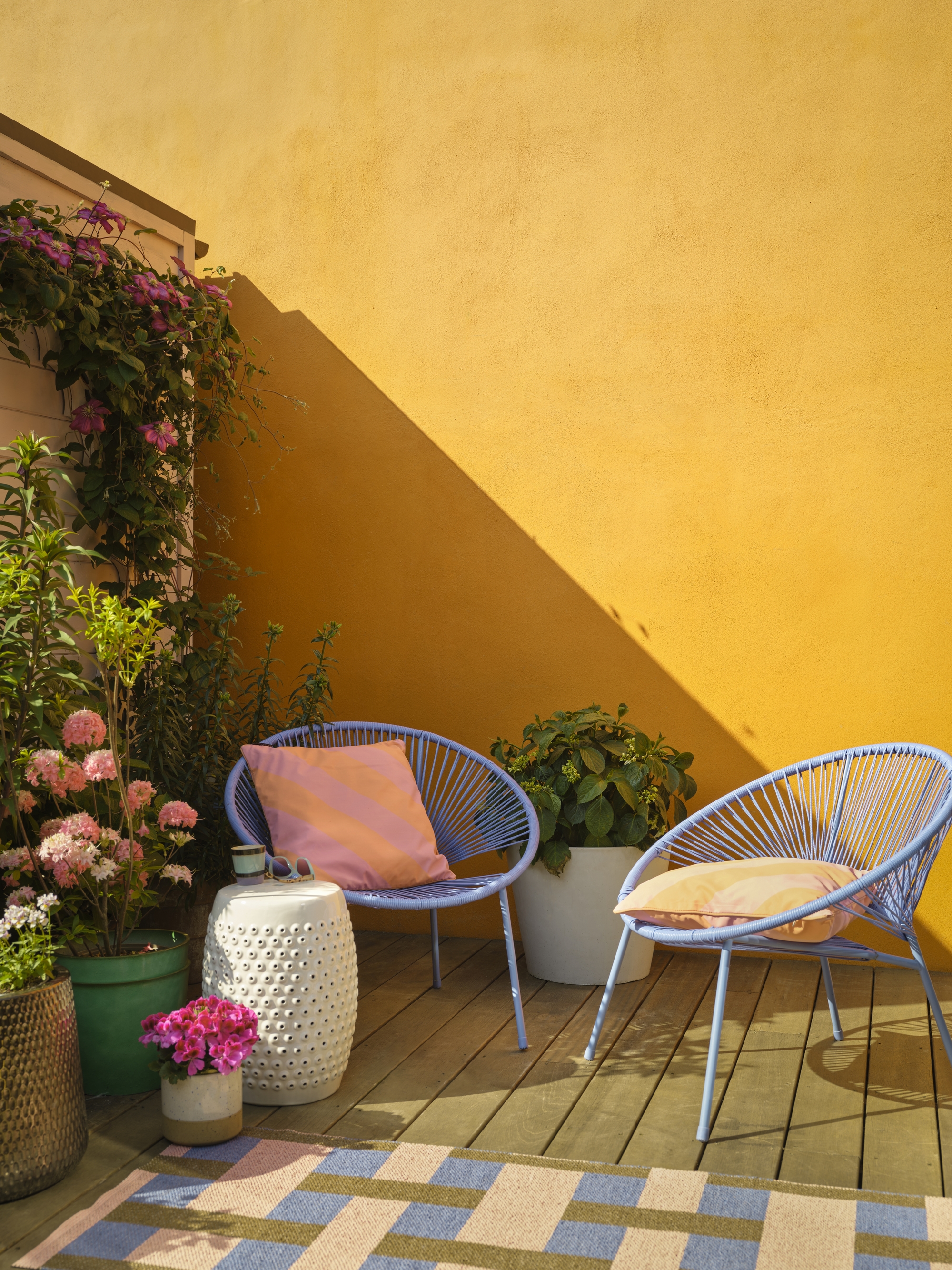 Image of an outdoor patio with bright yellow walls and lots of plants in the corner. There are two powder blue accent chairs with yellow pillows on them.