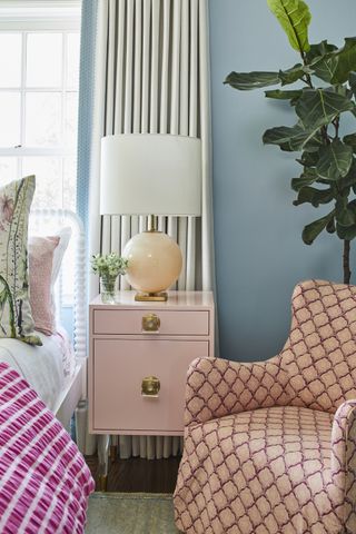 small bedroom with blue walls, pink nightstand, coral print armchair, bright pink blanket, plant, floral cushion, coral globe based table lamp with white shade, white drapes