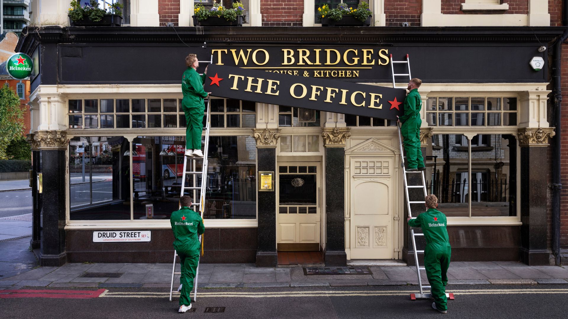 an image of a pub exterior having a new sign placed over their current one by a team of removal men in green Heineken overalls