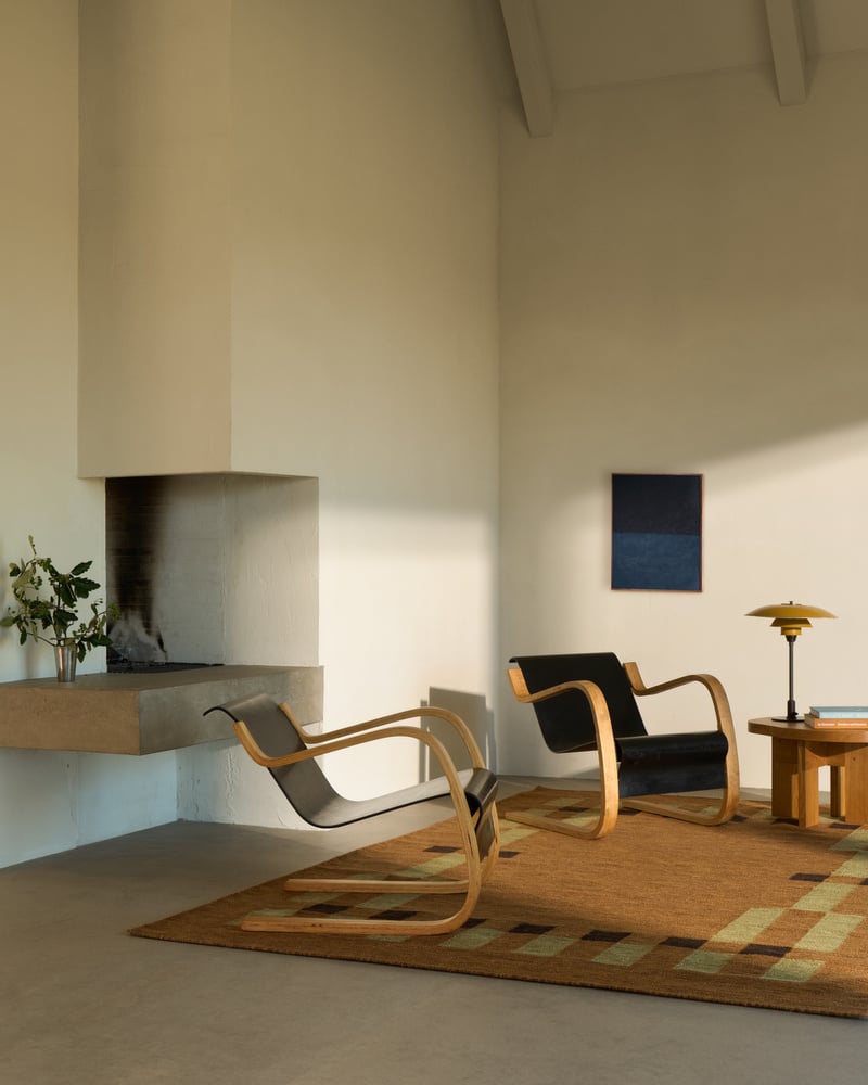 Image of a Scandinavian-style living room with white walls and a concrete floor. There is a tan flatweave rug with two modern accent chairs on it and a wooden side table.