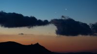 Two star-like planets shine close to one another in the evening sky above the silhouettes of clouds.