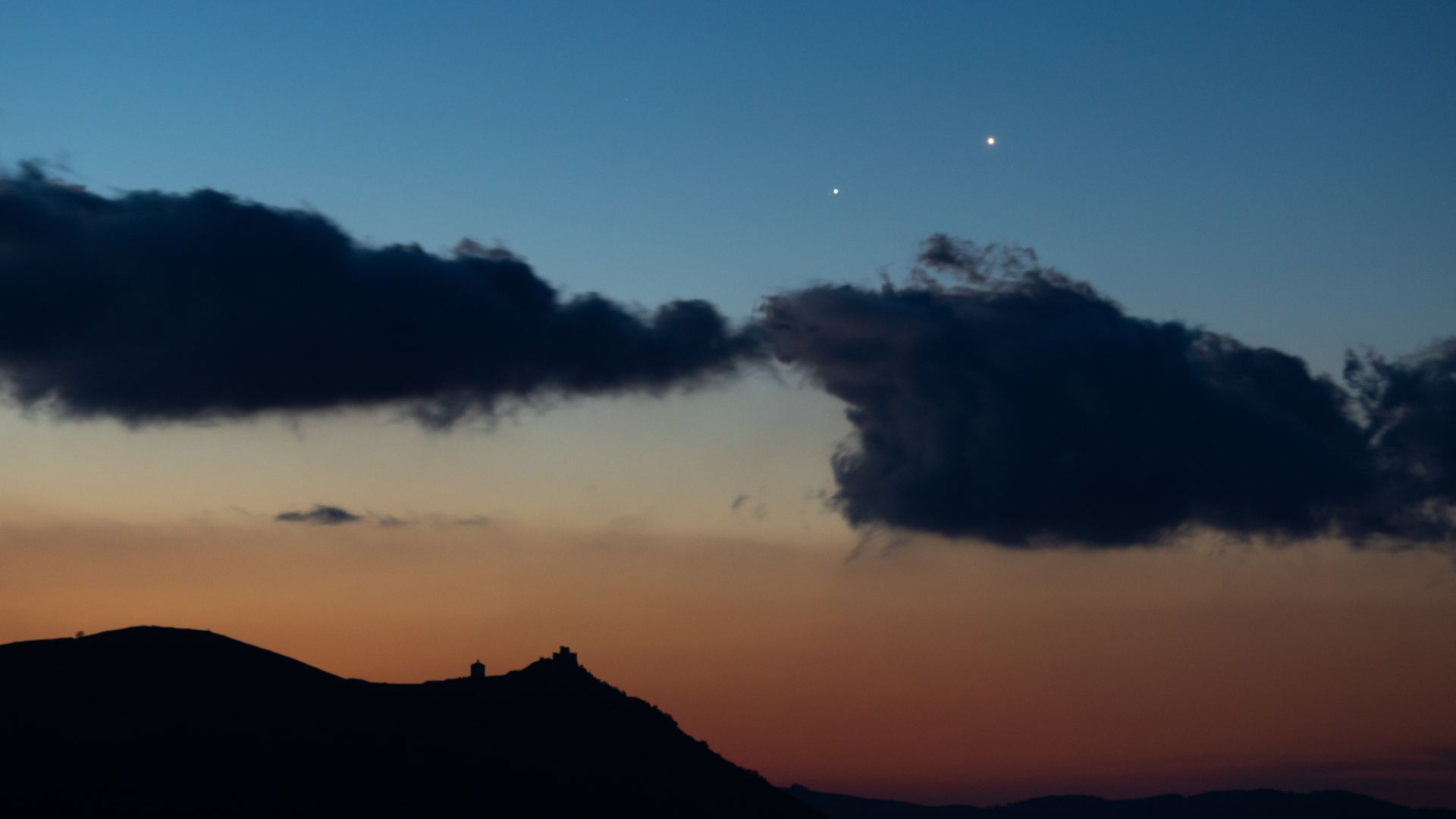 Two star-like planets shine close to one another in the evening sky above the silhouettes of clouds.