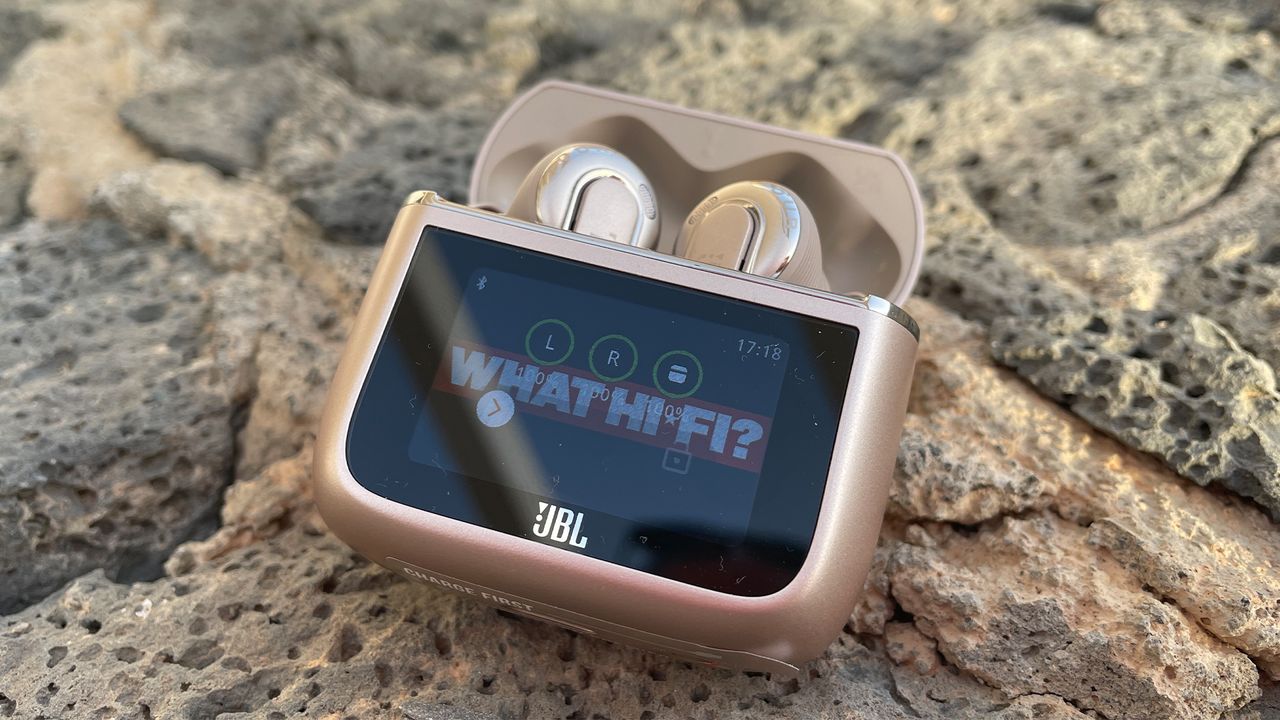 JBL Tour Pro 3 earbuds pictured on some rocks. The case is open and the buds are visible inside. The screen on the front of the case shows the What Hi-Fi? logo.