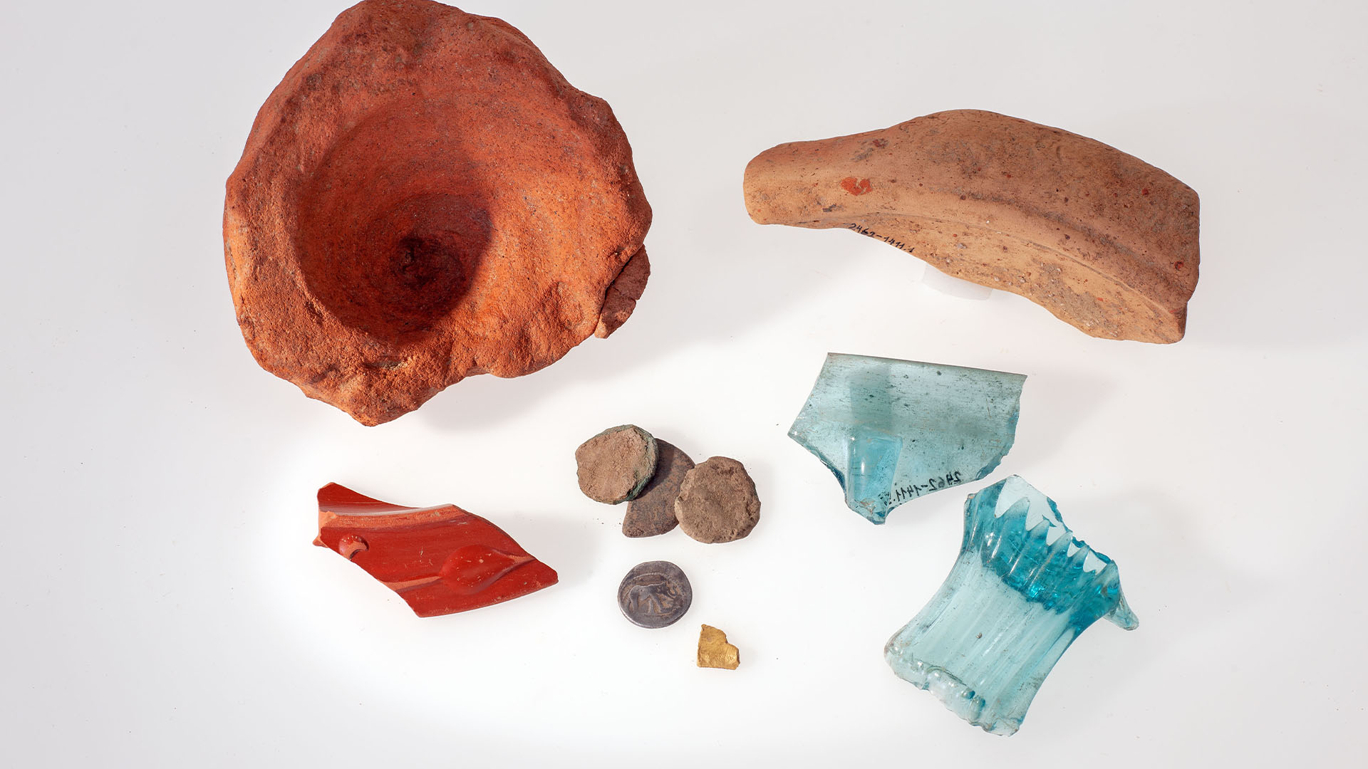 An amphora base, piece of a mortar, rim of a bowl, four coins, a gold object, pieces of a bottle and a blue glass bowl.