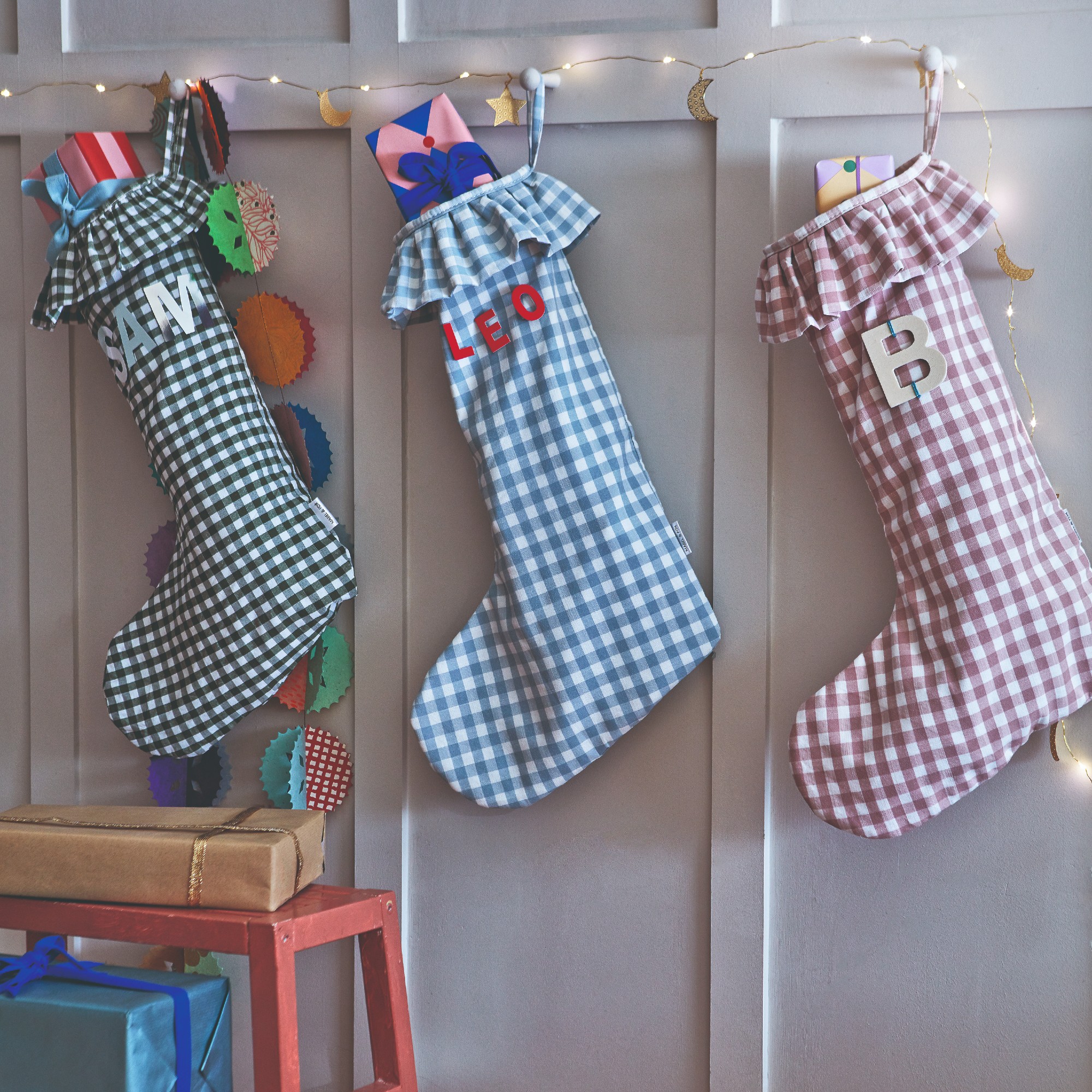 Three gingham ruffled Christmas stockings hanging on a panelled wall with moon-adorned Christmas lights surrounding them