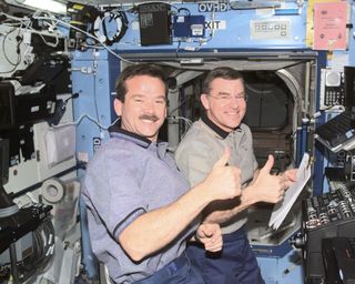 Astronaut Chris Hadfield on a space mission, front left, with a colleague back right. They are both floating in zero gravity and giving a thumbs up to the camera. They are surrounded by equipment, buttons monitors and wiring