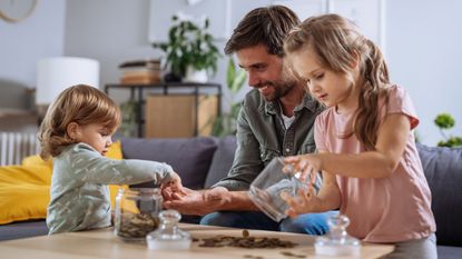 A dad and his daughters smile while putting coins in clear jars.