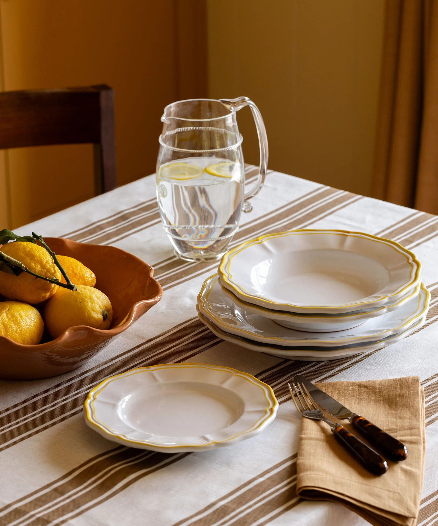 A table dressed with striped tablecloth, white plates with yellow rim, linen napkin, tortoiseshell cutlery, glass carafe, and terracotta bowl filled with lemons.