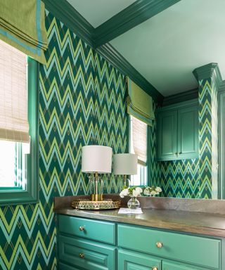 Green bathroom with green zig zag wallpaper, green painted ceiling and dark green ceiling trim, green cabinet with dark brown stone top, table lamp with white shade