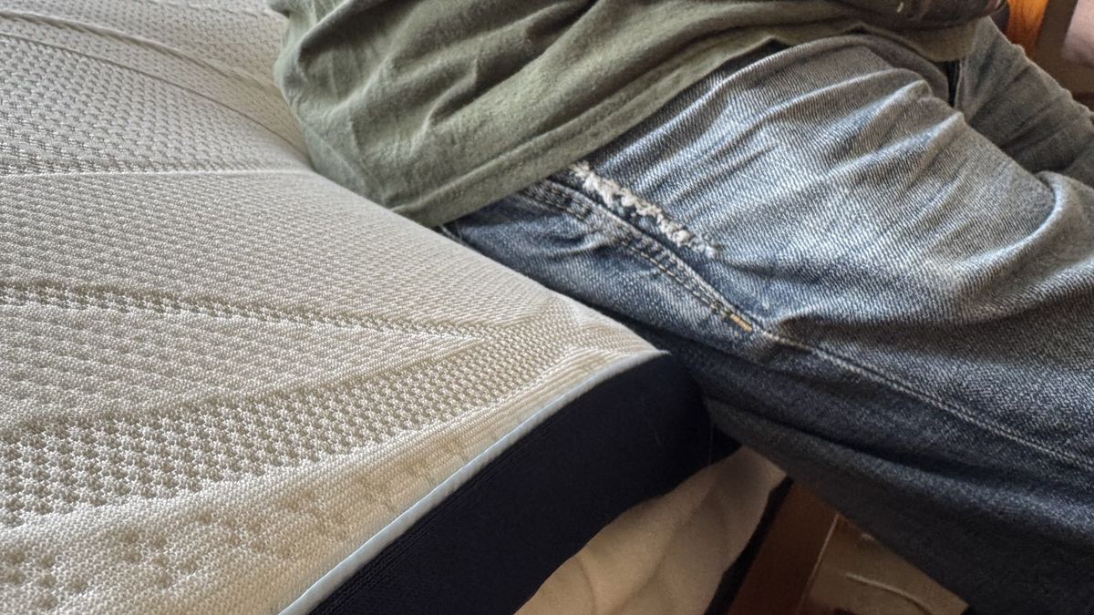 A close-up of person wearing blue jeans and a green t-shirt sitting on the side of the Nectar mattress topper.