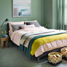 a green bedroom with green painted walls and a bed with pink bedn linen with two baskets on the floor at the end of the bed
