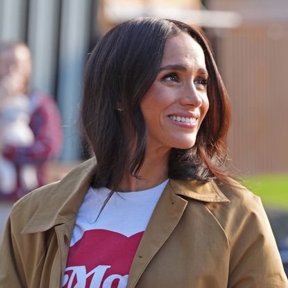 Meghan Markle smiling wearing a white and red t-shirt and a trench coat