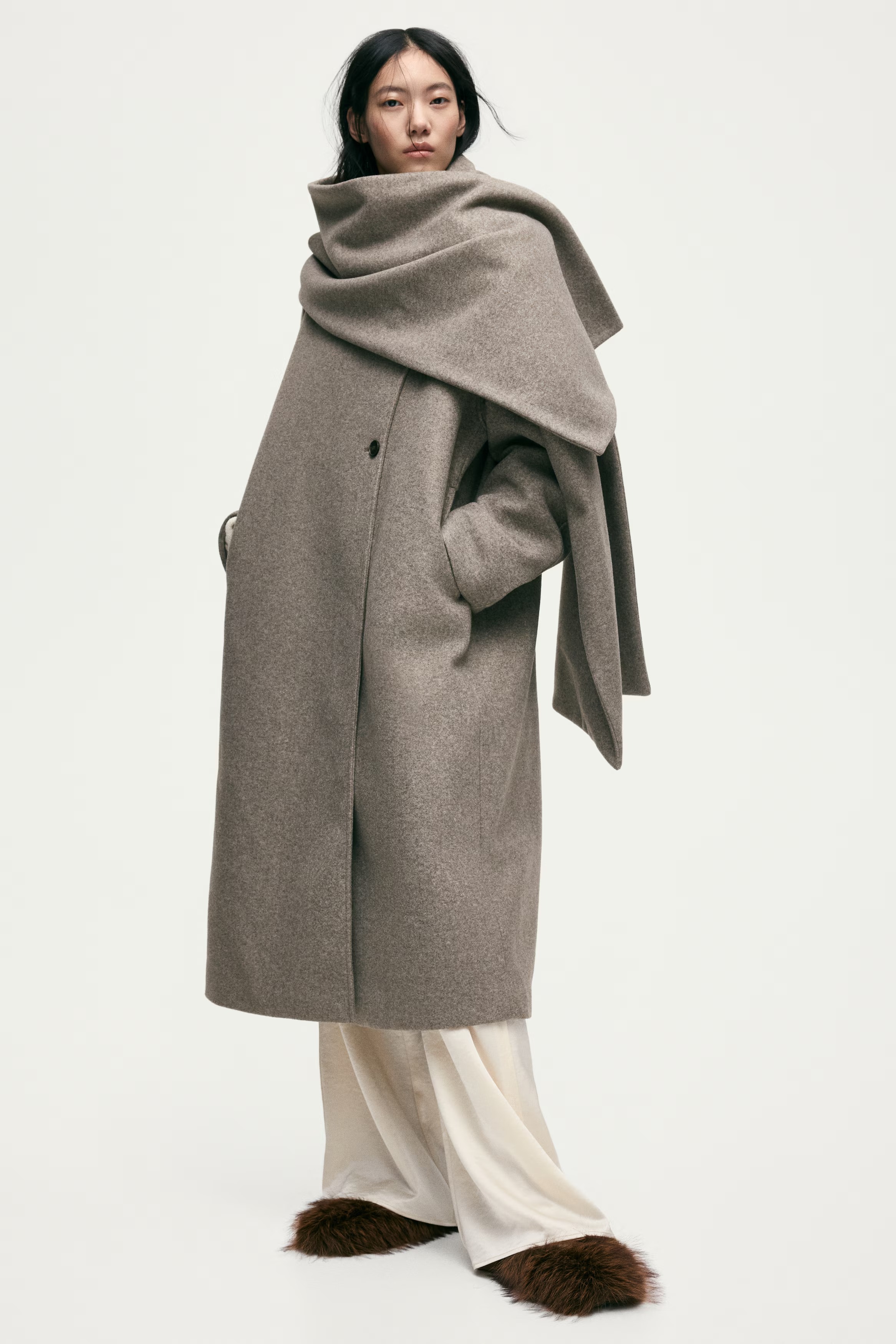 A model wearing a beige knee-length, double-breasted coat with a large, detachable scarf, round neckline, and a mixture of concealed and visible buttons at front.