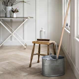 A metal bucket and mop on the floor of a room with a wooden stool and ironing board