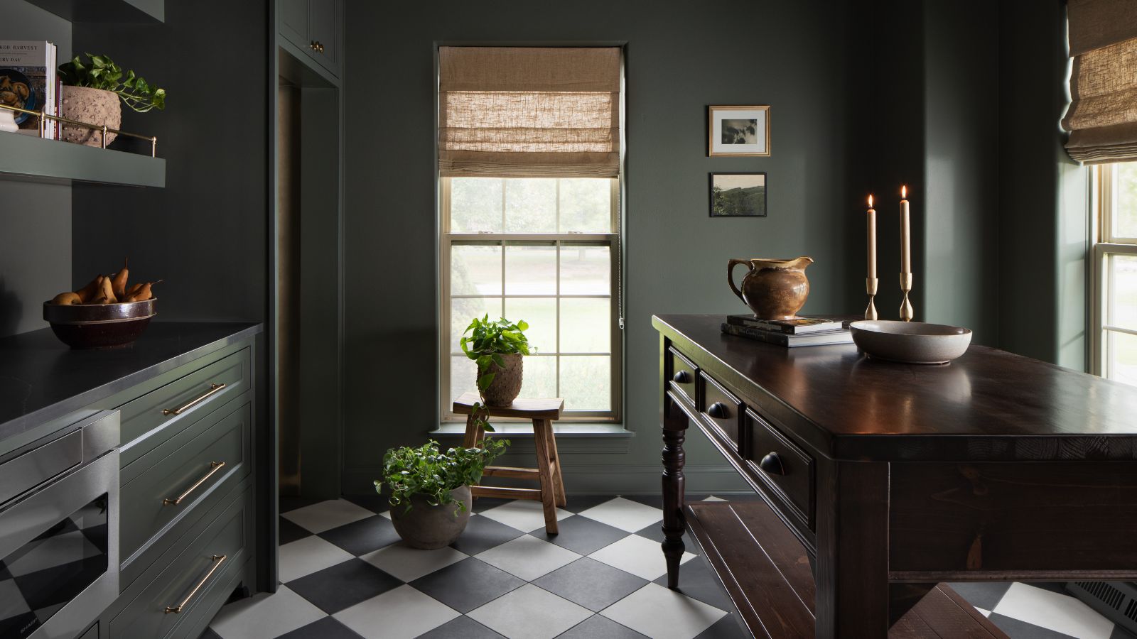 Dark kitchen area with dark brown wooden table and dark green cabinets, with bowls, candles and miscellaneous ornaments on countertops. Two windows reveal a bright day outside.