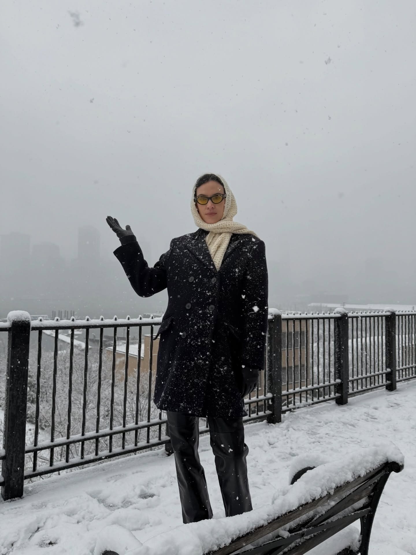 Alexa Chung stands outside in the snow wearing a black coat with a cream scarf.