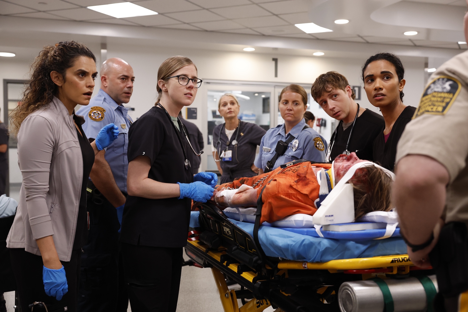 Sepideh Moafi, Taylor Dearden, Katherine LaNasa, Gerran Howell, Supriya Ganesh as doctors standing around a patient on a gurney talking to an officer in the pitt season 2