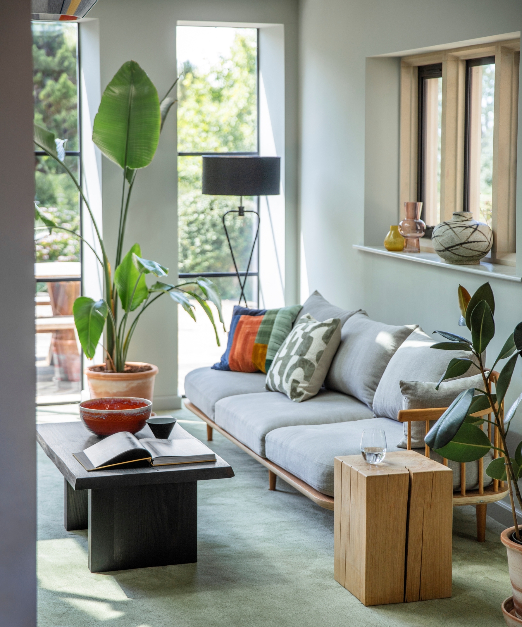Grey sofa with colourful cushions, wooden side table, tall plants, vases on windowsill in bright room.
