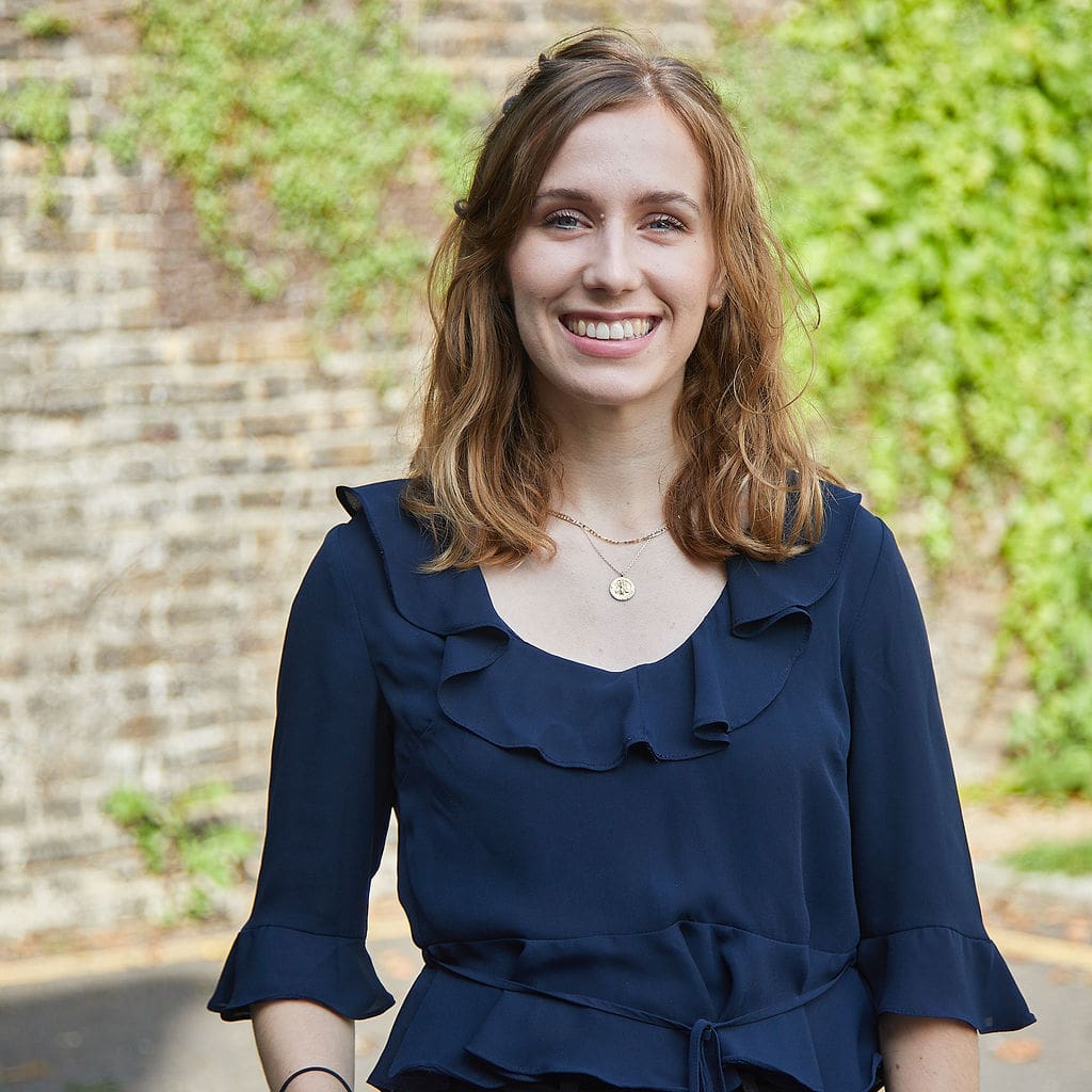 female with shoulder length light brown hair wearing navy top stood outside near stone wall with green trailing plants