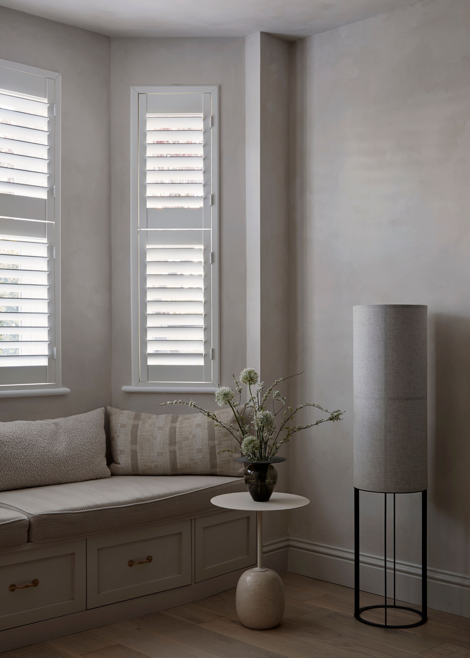 A corner of a living room area with seating framed around the curved window. The seating area also has extra storage. Beside the bench is also a side table with a vase and flowers on top as well as a floor lamp.