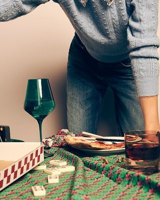 dark green wine glass sits on a floral tablecloth with other tableware and a woman leaning over