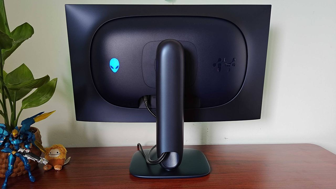 Back of Alienware AW2725Q gaming monitor with RGB logo on left sitting on woodgrain desk.