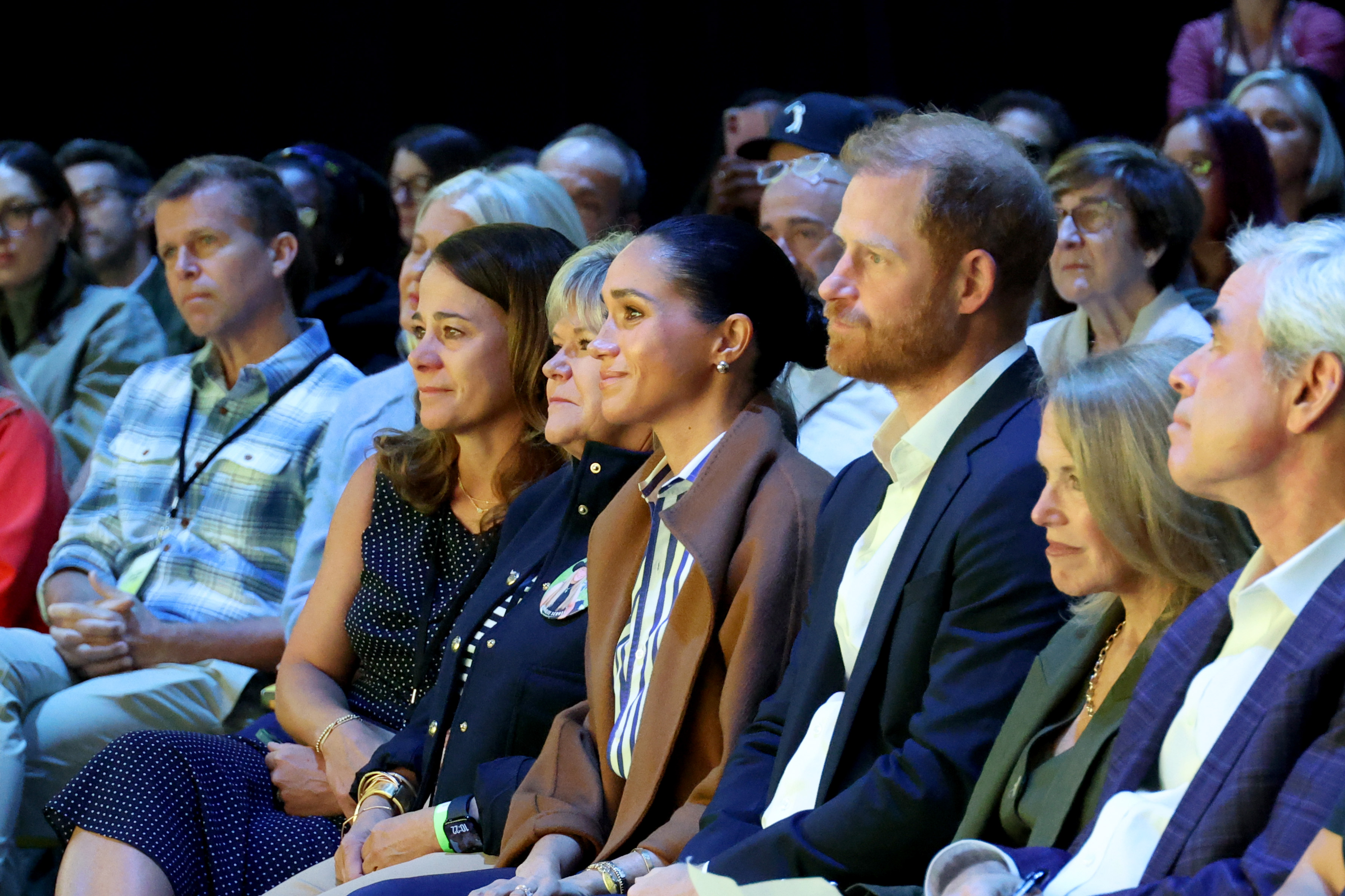 Meghan Markle wearing a brown blazer sitting next to Prince Harry in an audience