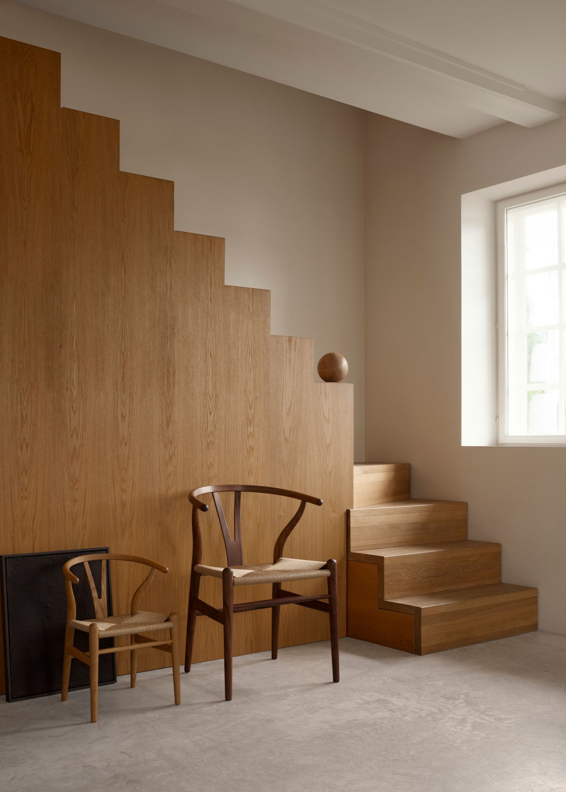 Two wishbone chairs beside each other in front of a wooden staircase in a white, minimalist room.