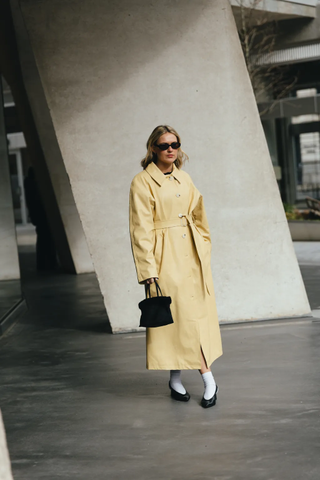 Fashion week attendee wears butter yellow trench coat, white socks and black shoes.