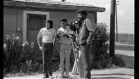 Four people outside a rustic building, setting up and operating a vintage film camera on a tripod in a rural area.