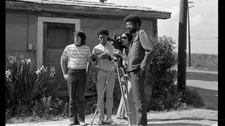 Four people outside a rustic building, setting up and operating a vintage film camera on a tripod in a rural area.