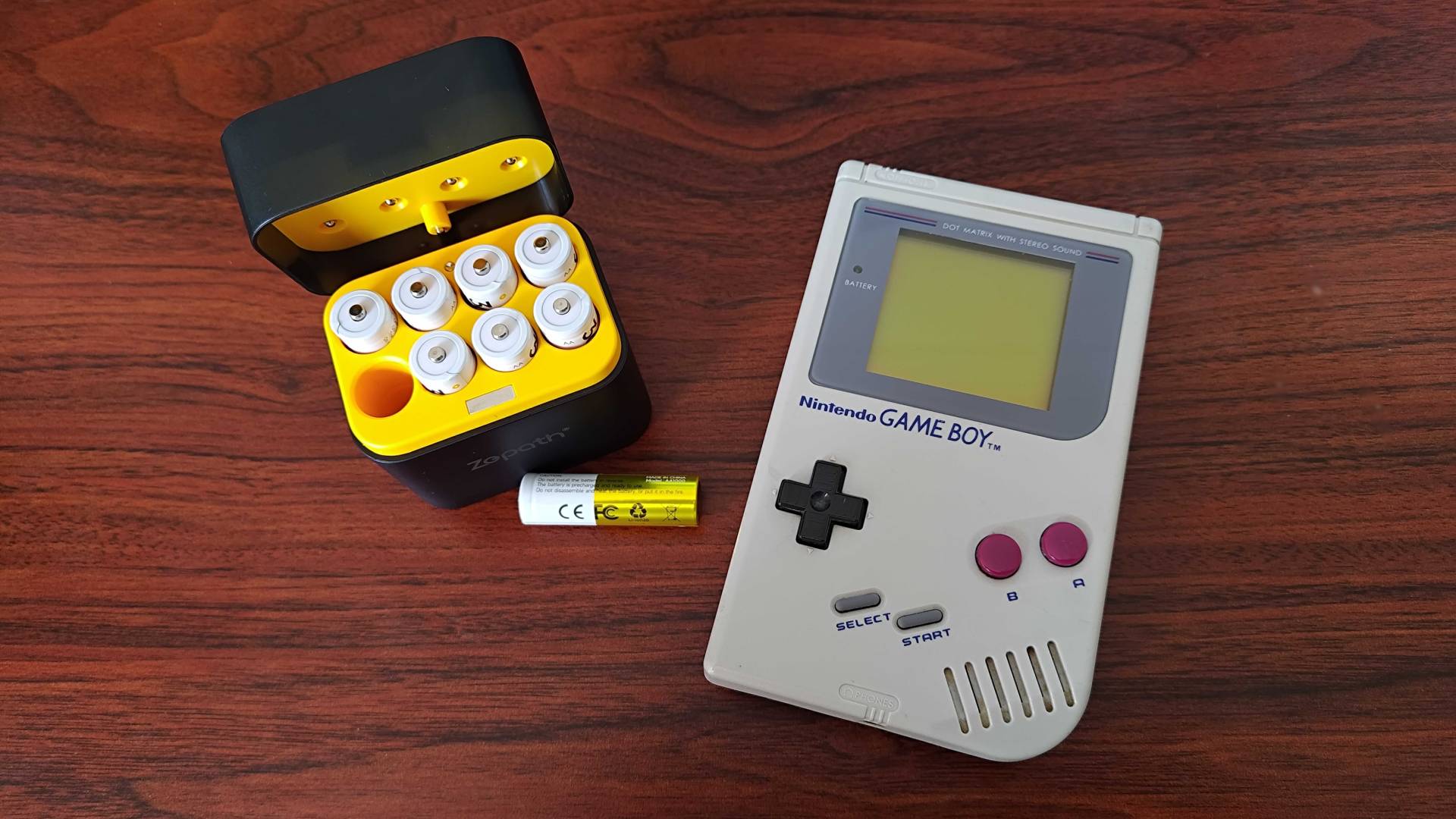 Rechargeable AA batteries with yellow and black charger next to original Game Boy on woodgrain desk.