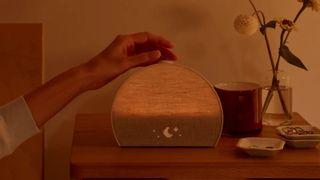 The Hatch Restore 3 alarm clock slightly lit up on a bedside table with a hand pressing a button on top of it