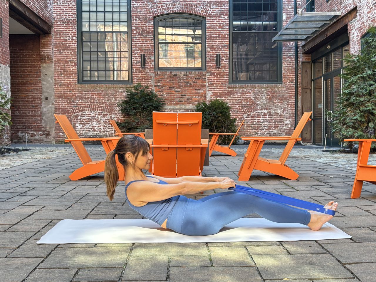 Pilates instructor Monica Borodin performs a half roll down. She wears a vest and leggings and is exercising outside on a mat. Behind her we see a brick building with large windows and table and chairs. She is seated, with her knees slightly bent and her legs virtually straight on the floor. She has looped a resistance band around her feet and holds it as she leans back.