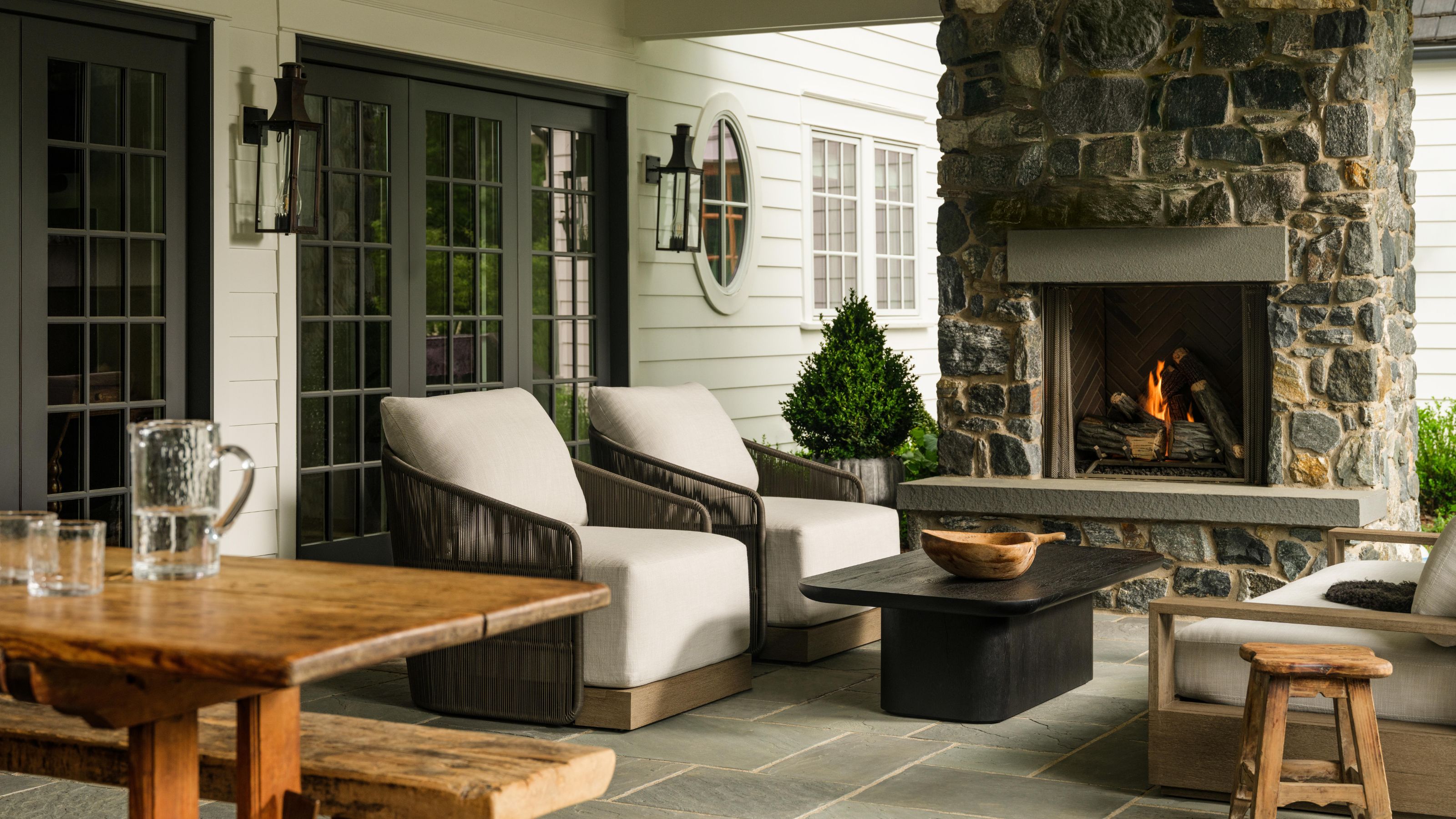 a covered porch off a historic cottage with a large stone al fresco fireplace, sitting area with coffee table and a rustic bench and table