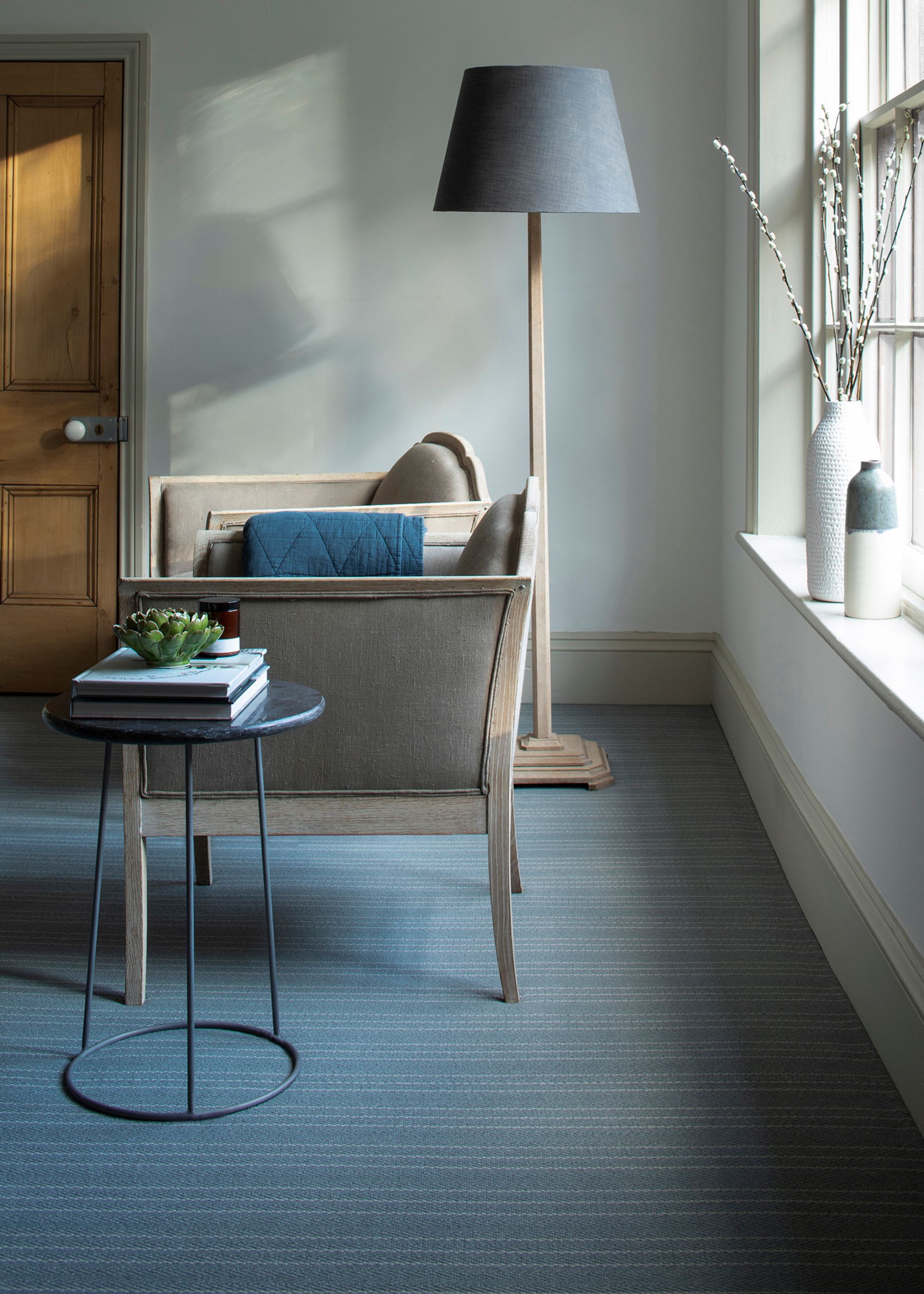 A grey floor covering with chairs and a side table. On the side table is a stack of books and a plant on top. In the corner of a room is tall wood lamp with a grey lampshade.