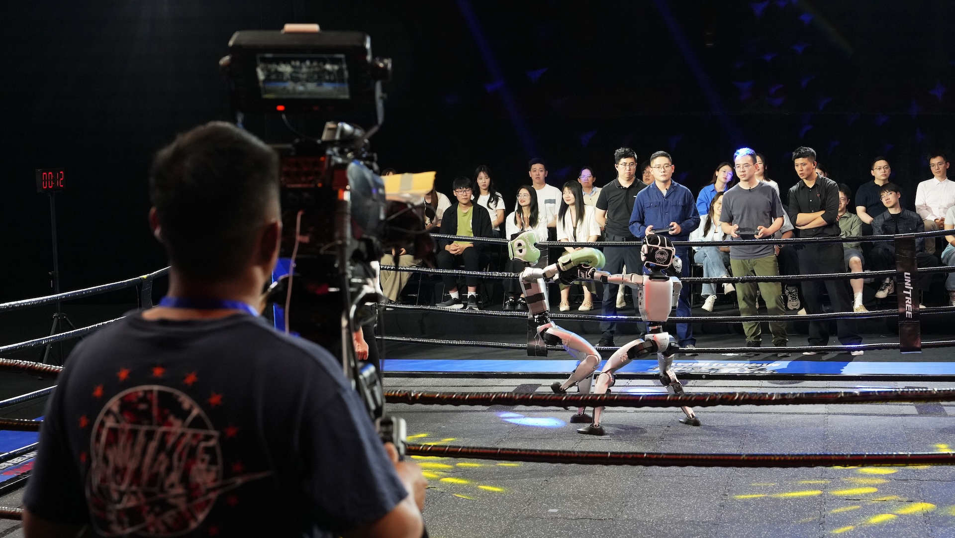 A cameraman records two robots fighting in a boxing ring as a crowd watches