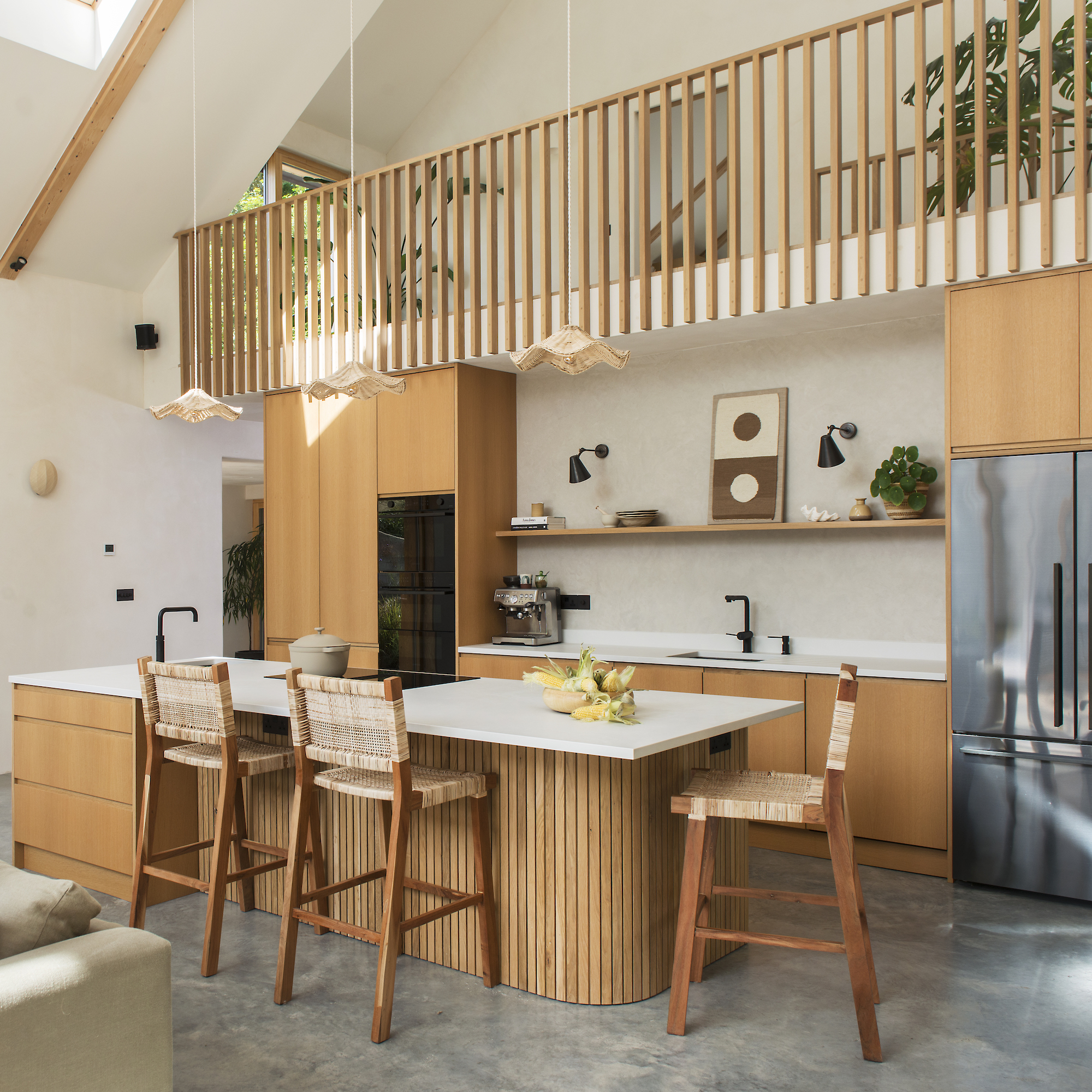 contemporary oak kitchen with wooden island with white counter, with hob and sink. wooden wall units, large metal refrigerator and mezzanine above.