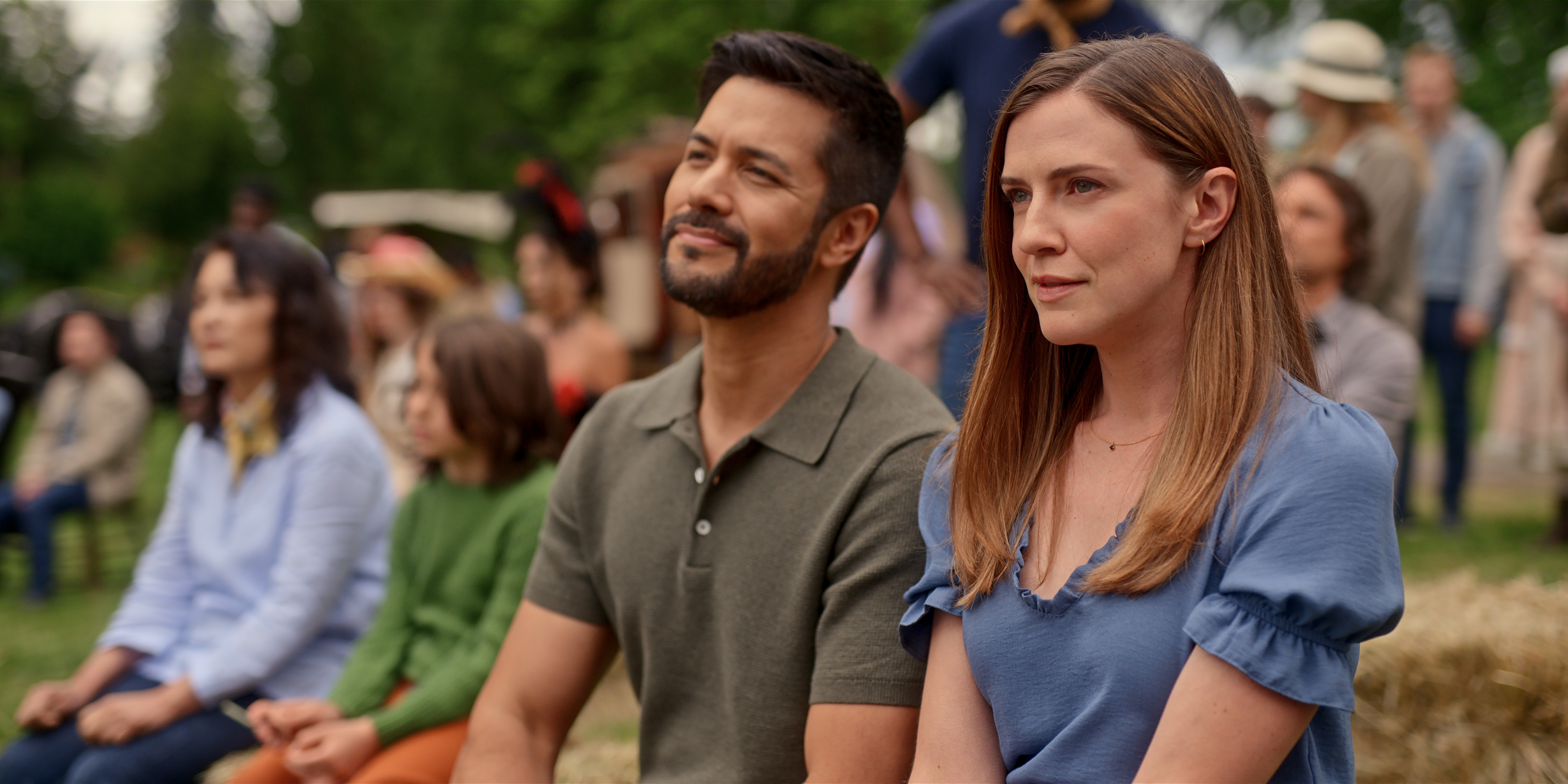 Marco Grazzini as Mike Valenzuela and Sara Canning as Victoria sitting on a hillside with a crowd in a still from virgin river season 7