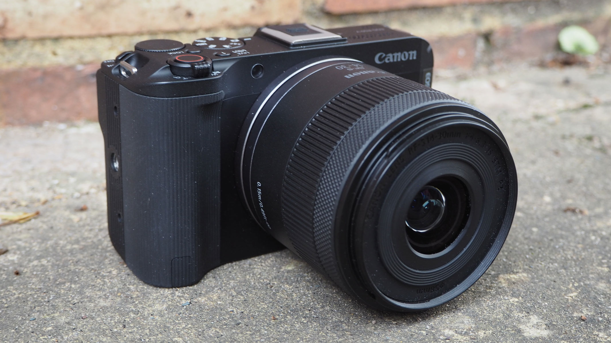 The Canon EOS R50 V on a concrete floor with bricks behind it.