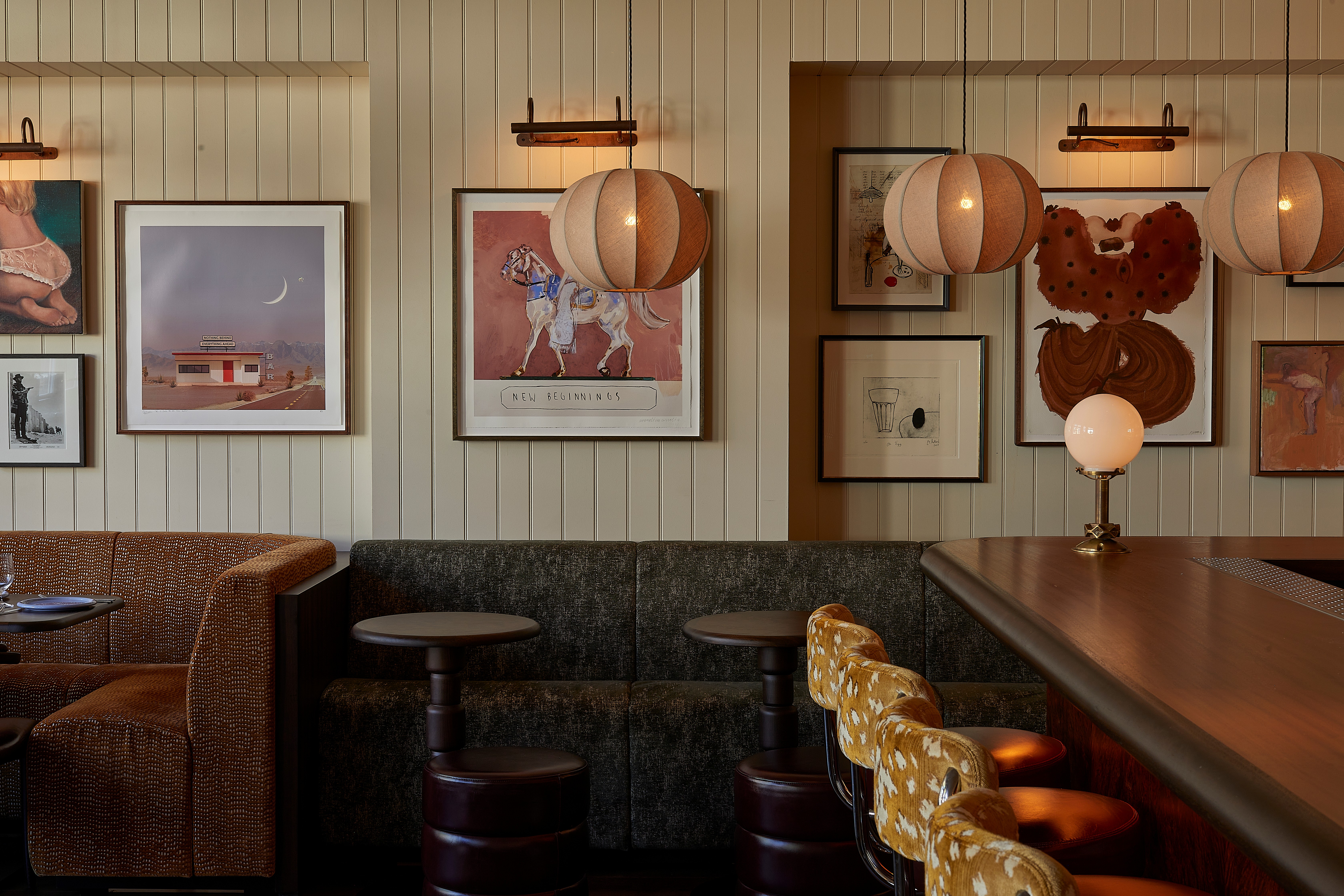 A Western-inspired dining room and bar with beige panelled walls, framed art, pendant lighting, and wooden furniture alongside upholstered stools.