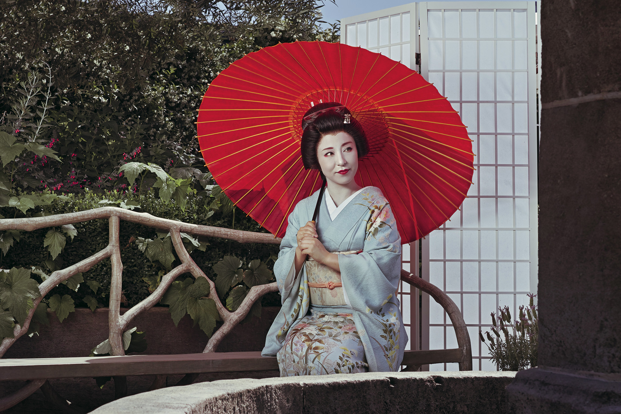 A woman in traditional Japanese attire sits gracefully under a vibrant red parasol, surrounded by lush greenery and a serene backdrop