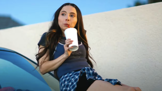 A still from the movie Drugstore June showing Esther Povitsky as June laid on a car and drinking something out of a big cup