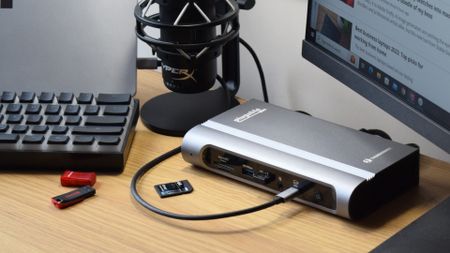 Plugable Thunderbolt 4 & USB4 Quad Display Docking Station (TBT4-UDZ) on a wooden desk with an SD card and USB drives in front of it.