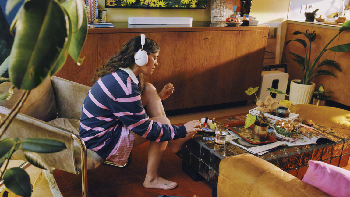 A person sat in a living room listening to Sonos Ace Ultra headphones with a Sonos Beam Ultra soundbar in the background