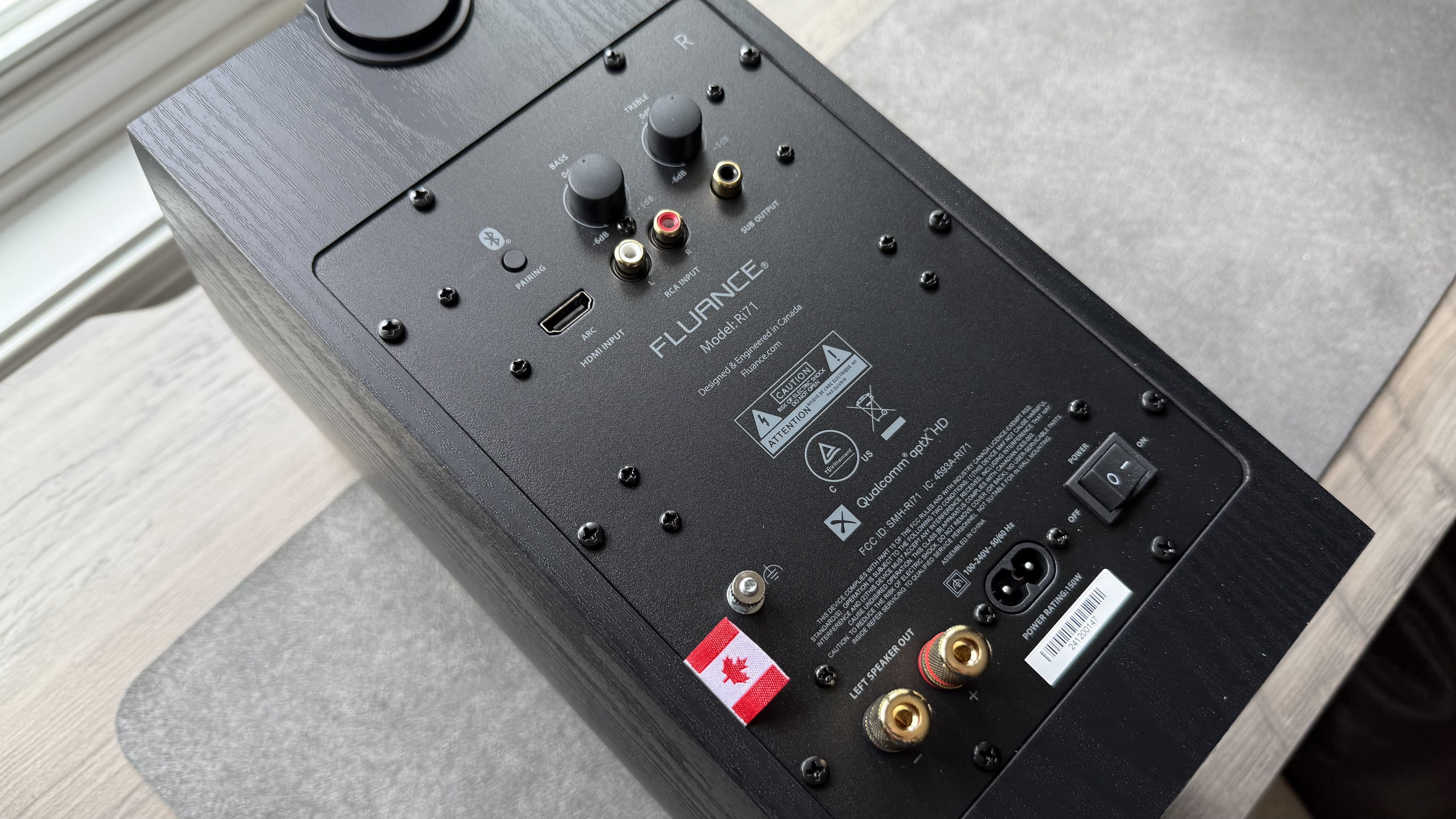 Rear panel of the Fluance Ri71 powered bookshelf speaker, placed facing upwards and showing hte ports and controls on the back, on a matt on a wooden surface.
