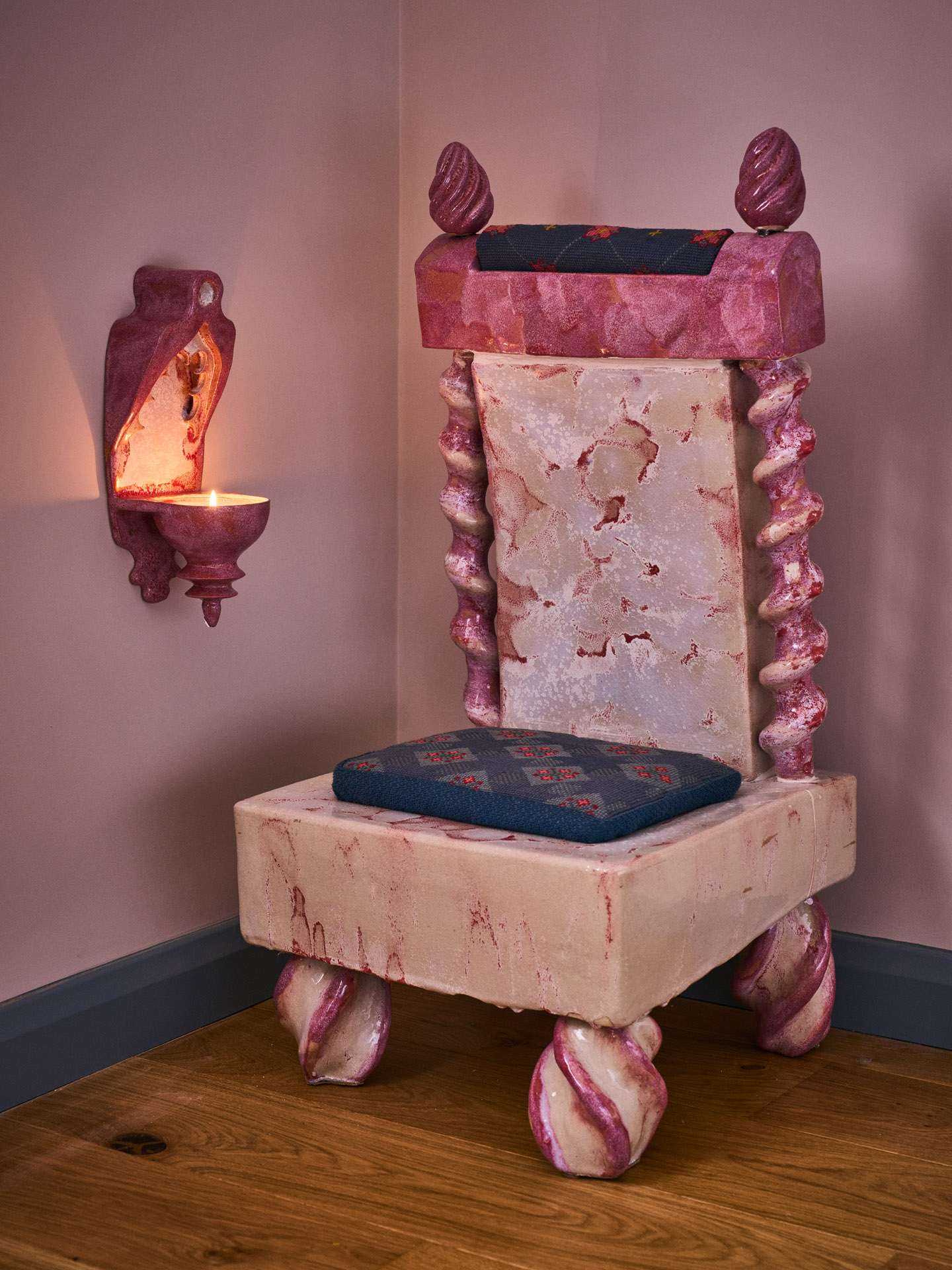 A pink armchair cast from ceramic and textiles in gradients of pink with inserted blue and red fabrics sits next to a candle sconce in the same style.
