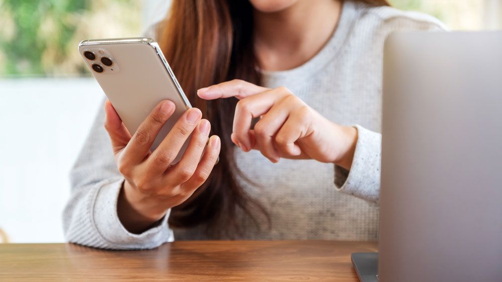 A woman using an iPhone with a MacBook open next to her.