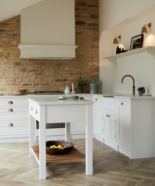 A white kitchen with a small kitchen island with an open wooden shelf, white kitchen cabinets around it, and a brown brick wall with a white wood hanging above the silver oven
