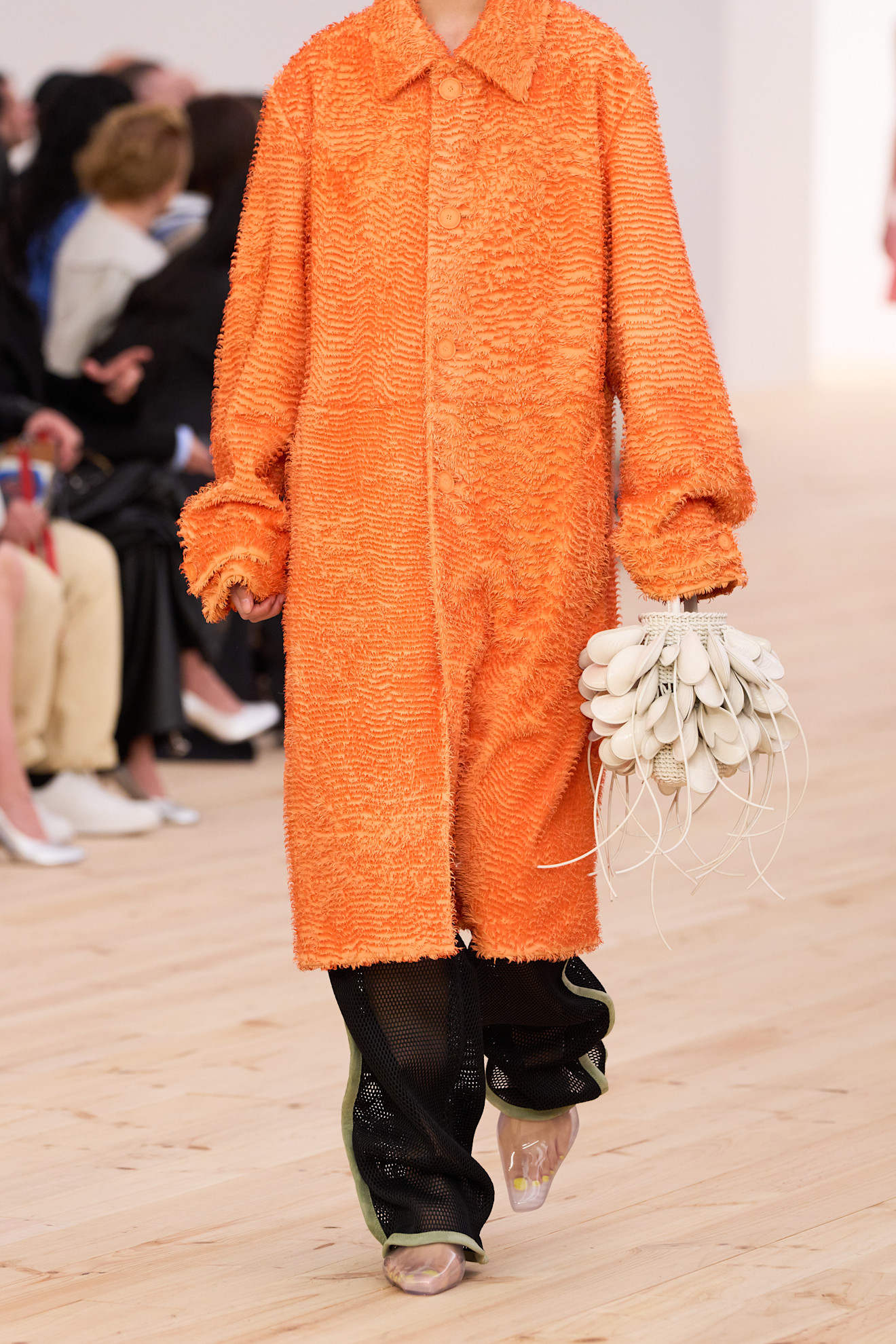 a model wearing a textured orange coat with a white clam bag and clear shoes on the Loewe Spring 2026 runway